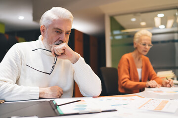 Senior businessman deeply contemplating documents at desk. businesswoman in orange cardigan diligently working in background. Concept of workplace focus, strategy, and professional dedication..