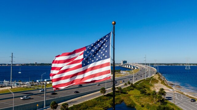Large American flag waving over a coastal highway with a bridge and blue sky in the background - Powered by Adobe