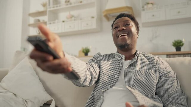 Portrait of laughing African man watching comedy by TV, sitting alone on sofa. Relax time in evening or weekend at home, entertainment and leisure, black person enjoying TV shows, cable television