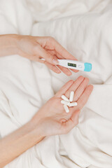 Person holding thermometer and pills on white sheet in bed with pillows