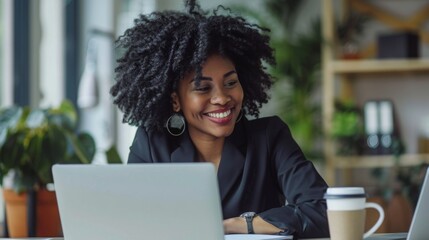 A woman with curly hair smiles while working on a laptop at her desk. A coffee cup sits to the right of her, and plants are visible in the background.