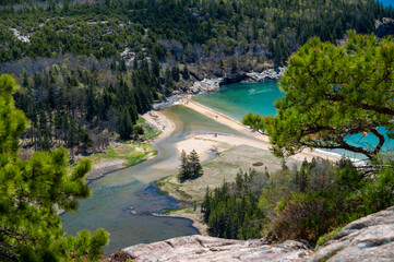 Aerial View of SandBeach and Ocean Inlet in Acadia National Park. A panoramic view from a clifftop overlooking a sandy spit connecting a rocky headland to a small, sandy beach. The clear blue water o