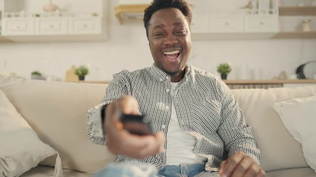 Portrait of cheerful African man watching TV and laughing, switching channels. Young black guy enjoying comedy and sitcom on television, leisure in weekend, spending time alone in apartment, pastime