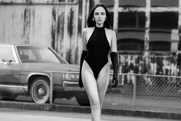 Fototapeta premium Sexy girl in bodysuit walking on the street. Hipster lifestyle, concept of brutal woman. Pretty stylish woman with legs spread posing near garage wall, urban city style. Sexy female body. Glamour