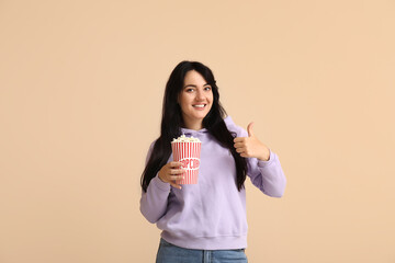 Happy young woman with bucket of popcorn showing thumb-up on beige background