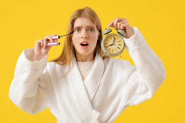 Hurrying young woman with alarm clock applying mascara on yellow background
