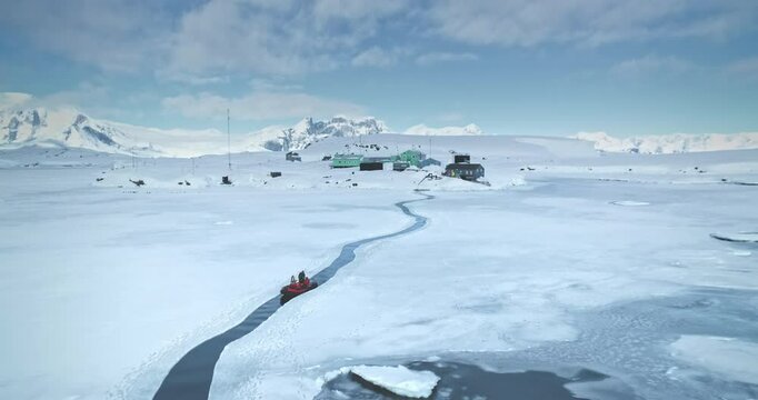 Scientist from polar station base riding zodiac boat in melted steam. Frozen ocean ice, mountains and science buildings in background. South Pole tourism, travel, expedition. Aerial drone panorama