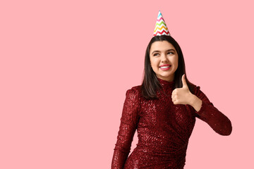 Beautiful young woman in party hat showing thumb-up gesture on pink background