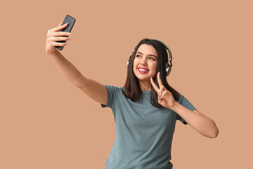 Beautiful young woman in modern headphones taking selfie and showing victory gesture on brown background