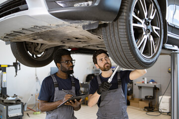 Focused automotive technicians working on car maintenance and safety inspection. Two mechanics check vehicle chassis on lift in professional auto repair shop.