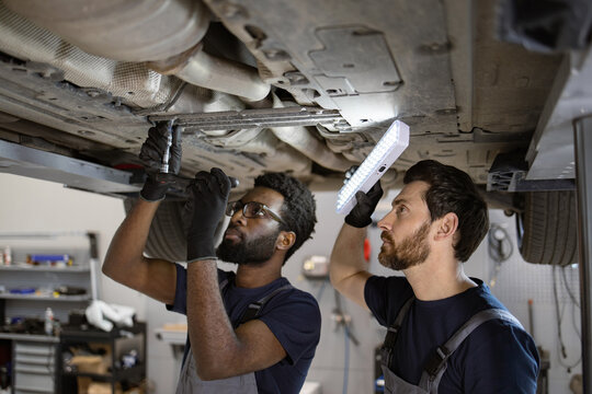 Two mechanics working diligently under car in auto repair shop. Focused technicians using tools and light to inspect and fix vehicle. Professional teamwork and precision in automotive maintenance.