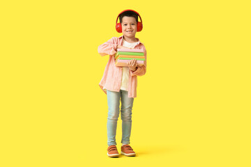 Cute little Asian boy in headphones listening to audiobook with books on yellow background
