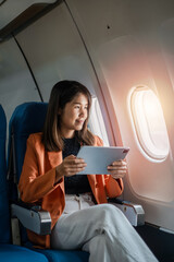 A young woman in a bright orange blazer uses a tablet while seated on an airplane, enjoying in-flight entertainment during her travel.