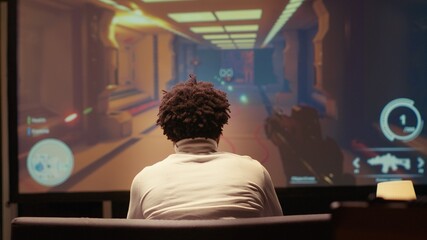 Man playing first person shooter videogame on smart TV in futuristic industrial setting with health, stamina, ammo bars HUD overlay. Gamer enjoying competitive PvP fps online multiplayer game