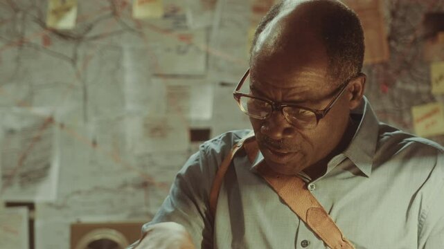 Serious African American investigator having discussion with witness examining mugshots of suspects during interview. Over the shoulder view
