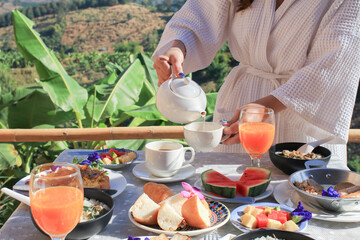 Woman hand pouring hot tea in white cup on wooden table set breakfast food outdoor in resort. Healthy food.