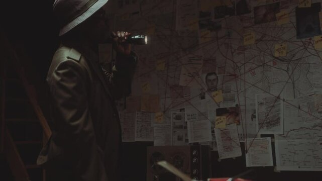African American detective wearing trench coat and fedora hat holding flashlight and studying detailed evidence board in dark office at night when investigating crime