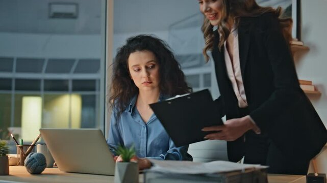 Corporate mentor collaborating intern at night desk. Smiling woman show document