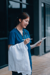 A cheerful woman in scrubs holds a clipboard, standing confidently before a modern hospital...