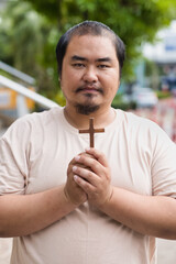 Middle-aged Asian Christian man showing wooden holy cross and praying with strong faith in Christianity