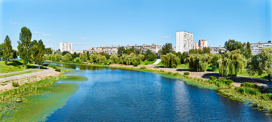 Fototapeta premium Scenic urban park with a calm river, lush green trees, and apartment buildings under a clear blue sky, creating a harmonious blend of nature and city life.