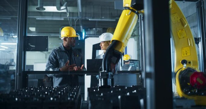 Two Industrial Engineers Working in a Facility, Black Male and a White Female in Hard Hats, Discussing the Automation Benefits of an Operational Robotic Arm in a Modern Technology Factory