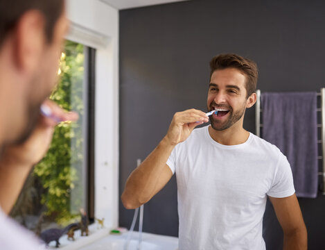 Man, smile and brushing teeth in mirror of bathroom for oral hygiene, fresh breath and morning routine in home. Happy person, reflection or cleaning mouth for dental health, self care or healthy gums