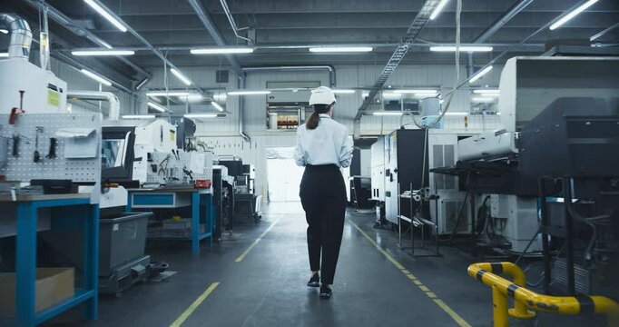Female Worker in a White Hard Hat Walks Through a Manufacturing Facility, Inspecting Machinery and Ensuring Operational Efficiency. Factory is Equipped with Advanced Automated Engineering Tools