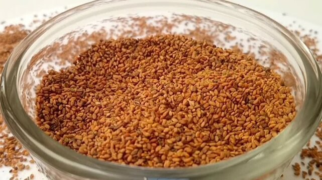 Fenugreek seeds drops in a rotating glass bowl on white background with spices spilled around it, micro shot, center frame, herbs and spices.