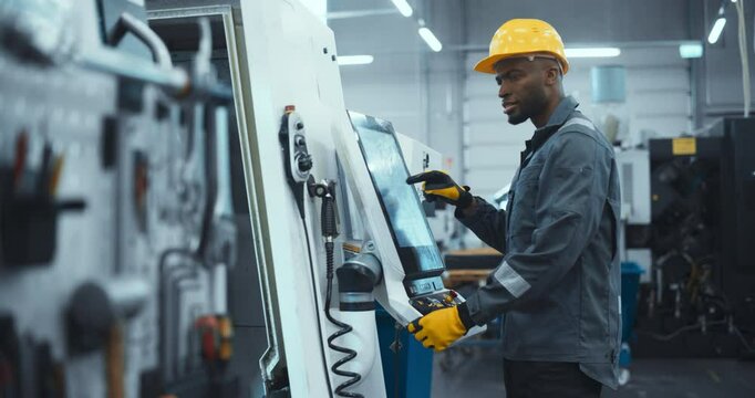 African Male Engineer in a Yellow Hard Hat Operating a CNC Milling Machine in a Modern Factory. Young Technician Inserting Parameters on a Touch Screen Display, Automating Production Line