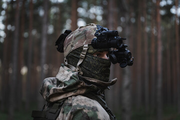 A soldier in balaclava and camouflage, with a night vision binocular on his head in the forest at dusk.