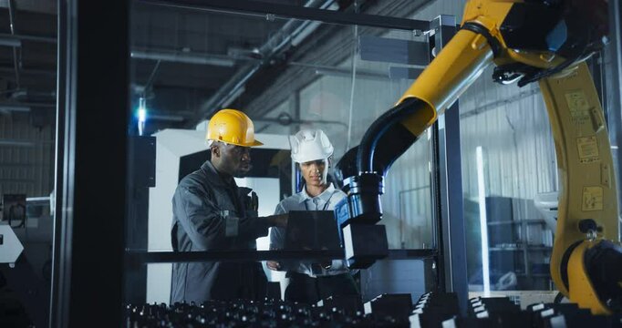 Industrial Manufacturing Facility: Young Black and Hispanic Engineers Controlling a Robotic Arm with a Tablet Computer App. Operators Downloading New Production Instructions for the Machine