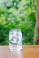 a glass of cold water with ice on wooden table in garden background.