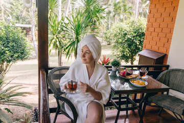 A woman in a white robe sips tea on a balcony with a view of lush greenery, enjoying a relaxed morning in a tropical setting