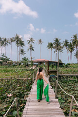 Woman in a green outfit walks along a wooden bridge over a lush green field of vegetation, surrounded by palm trees. The sky is blue with white clouds