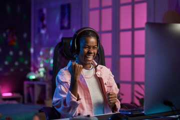 Portrait of young Black woman enjoying gaming night at home with purple neon lights and celebrating...