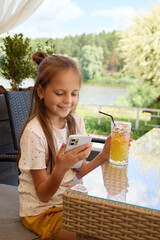 Charming smiling brown-haired Caucasian girl enjoying refreshing delicious drink at cafe and browsing kids content in internet enjoying online scrolling