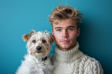 Young man with messy blonde hair is posing with a small white dog on a blue background, owner and dog look alike. 