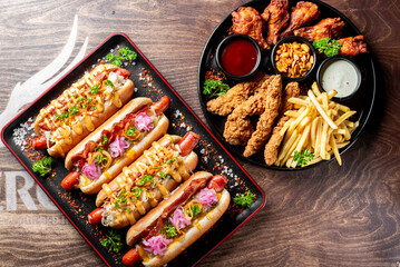 A vibrant food platter featuring gourmet hot dogs with various toppings, a side of crispy chicken tenders, golden french fries, and assorted dipping sauces on a rustic wooden table.