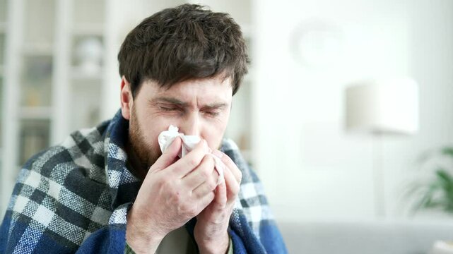 Sick man suffering from a cold wrapped in blanket sitting on sofa in living room at home. A tired bearded male coughs, sneezes, blows his nose with a handkerchief. He has fever or flu. Close up