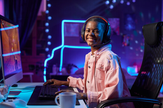 Side view portrait of joyful Black teenage girl playing video games or recording podcast with neon lights and looking at camera with smile copy space - Powered by Adobe
