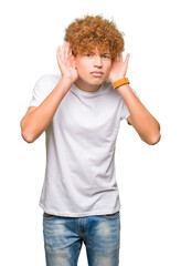 Young handsome man with afro hair wearing casual white t-shirt Trying to hear both hands on ear gesture, curious for gossip. Hearing problem, deaf