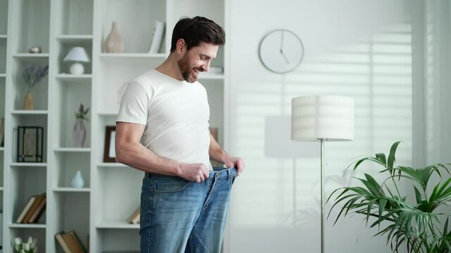Happy handsome man feels joy after losing weight. Smiling satisfied middle aged bearded male tries on his big pants, which he wore before, standing in living room at home looking at the camera