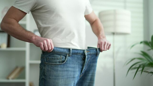 Close up of male hands trying on his big pants after losing weight. A middle aged man is happy with the success of a good figure waist after a diet while standing in living room at home
