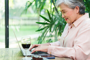 Elderly Asian woman in a blue denim shirt working on a laptop in a modern restaurant. Natural light from large windows, green plants outside, glass of wine on the wooden table. Focused on work,