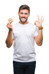 Young handsome man eating chocolate chips cookie over isolated background doing ok sign with fingers, excellent symbol