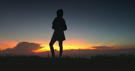 Fototapeta premium A silhouette of a woman standing on a hilltop, gazing out at a colorful sunset. The image was captured at a resort in Thailand, showcasing the beauty of the natural landscape.