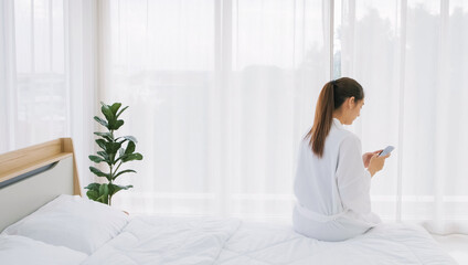 The back of long tied hair Woman in a bathrobe using a smartphone while sitting on the bed with a closed white curtain over the glass window in the background. Image with copy space.