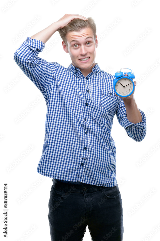 Wall mural young handsome blond man holding alarm clock stressed with hand on head, shocked with shame and surp