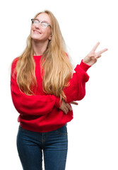 Blonde teenager woman wearing red sweater smiling with happy face winking at the camera doing victory sign. Number two.